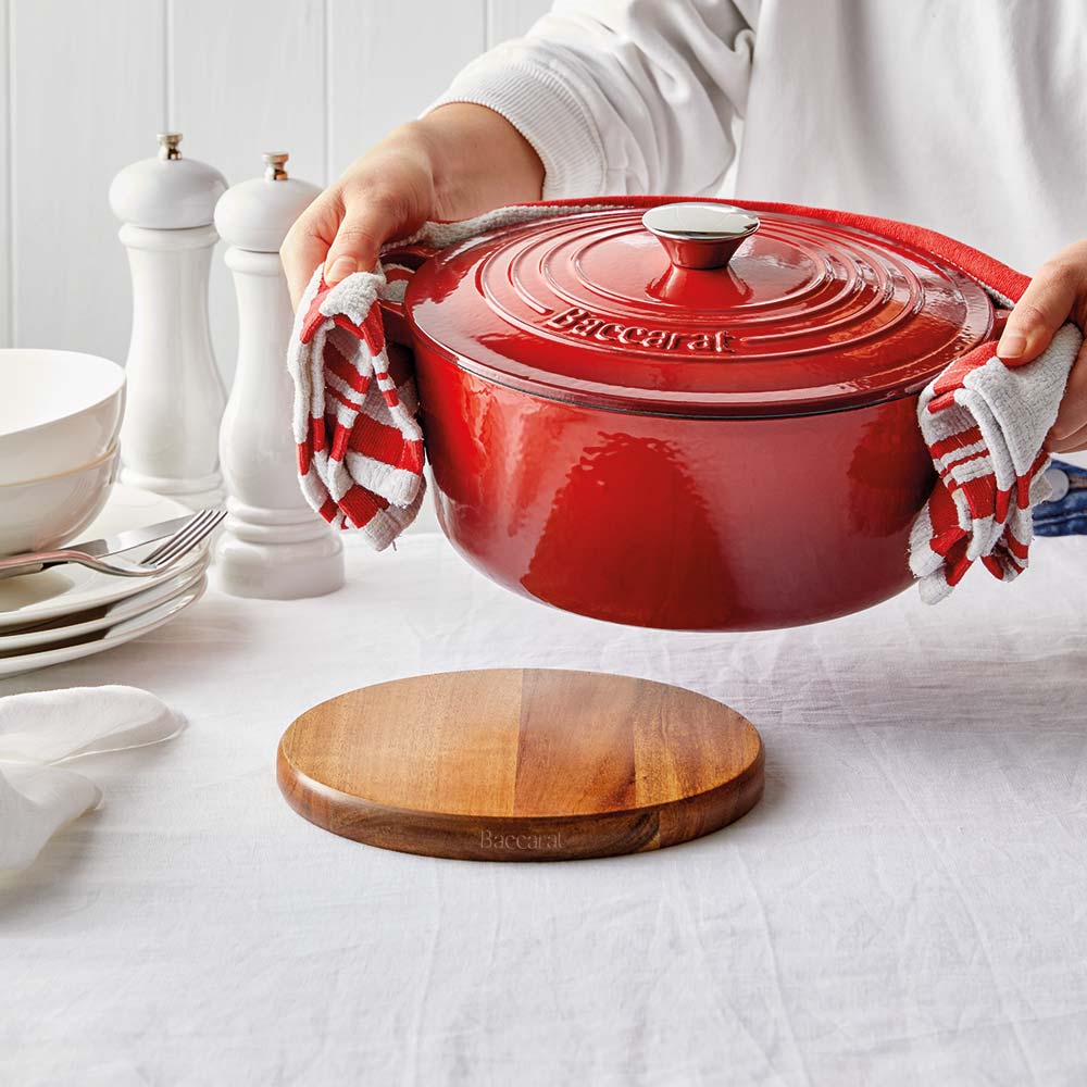 A hand gently holding a wooden trivet under a shiny red pot on a white surface red pot - Baccarat