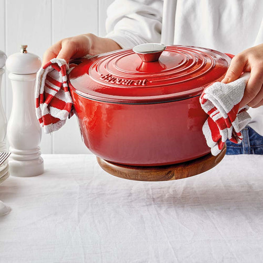 A hand gently holding a wooden trivet under a shiny red pot on a white surface red pot - Baccarat
