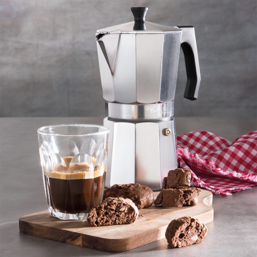 A silver Espresso Maker stands on a wooden cutting board next to a glass of espresso and chocolate cookies, with a red-and-white checkered cloth on a gray surface.