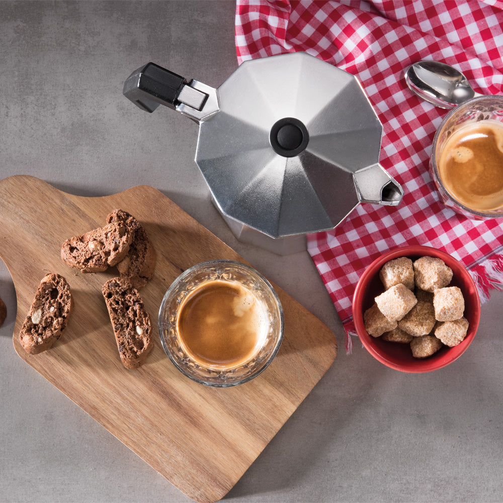 An overhead view of silver espresso-making equipment is arranged on a gray tabletop with a red-and-white checkered cloth, a glass of espresso, biscotti on a wooden board, and a bowl of sugar cubes.