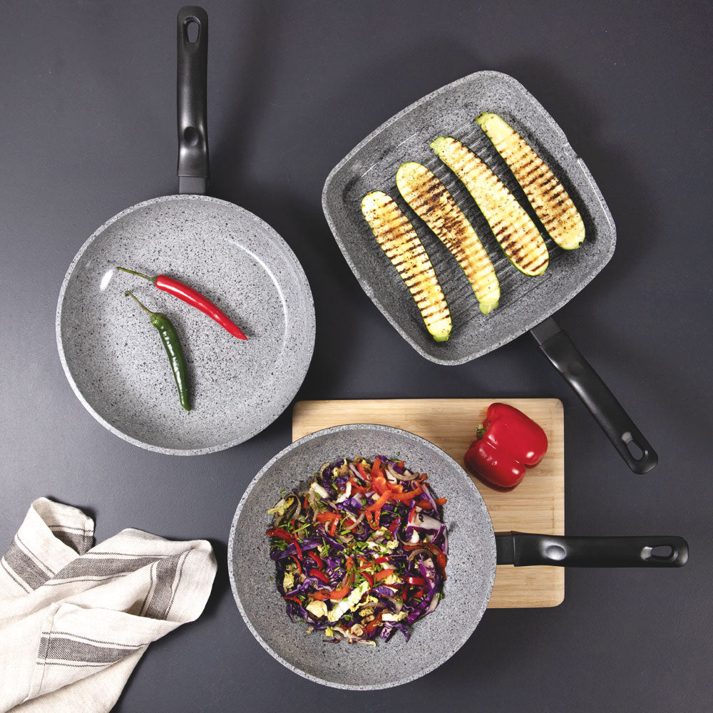 Three gray nonstick cookware pans arranged on a dark gray surface with food inside: one pan with two peppers, one square pan with grilled corn on a wooden board, and one pan with mixed vegetables. A folded striped cloth is visible at the lower left.