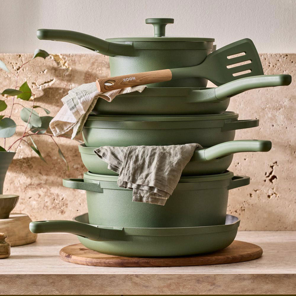 A stack of olive green cookware, including pots, pans, lids, and a spatula, arranged on a wooden board on a kitchen counter with a textured stone wall and a linen towel.