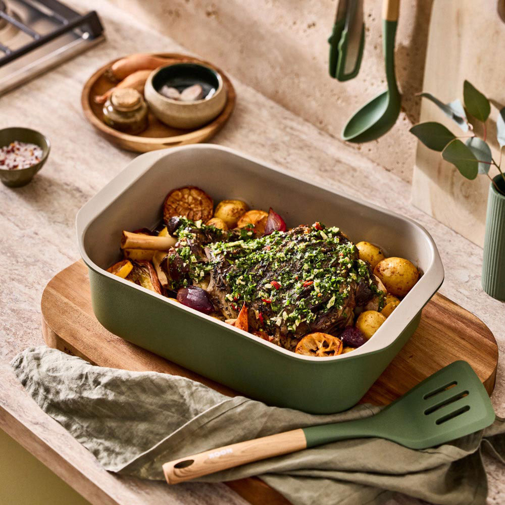 A roasted dish with vegetables is served inside a rectangular sage green pan on a wooden board in a warm kitchen setting.