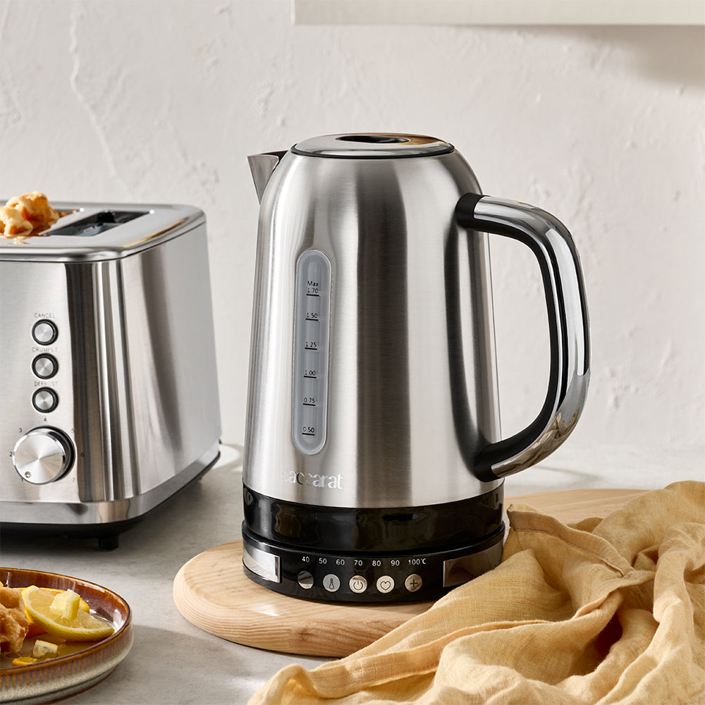 A stainless steel electric kettle with a black base and handle, standing on a wooden coaster next to a toaster and a beige cloth on a kitchen counter.