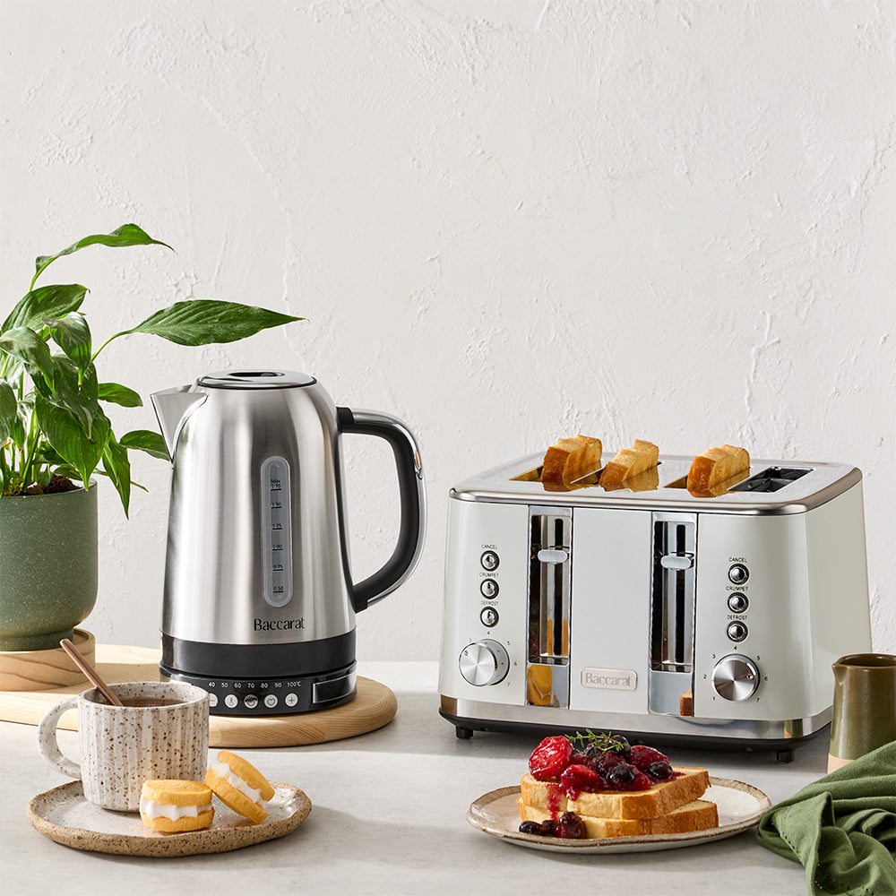 A stainless steel electric kettle and a cream two-slot toaster with toast inside, styled on a countertop with a plant, a mug, and a plate of breakfast items against a textured off-white wall.