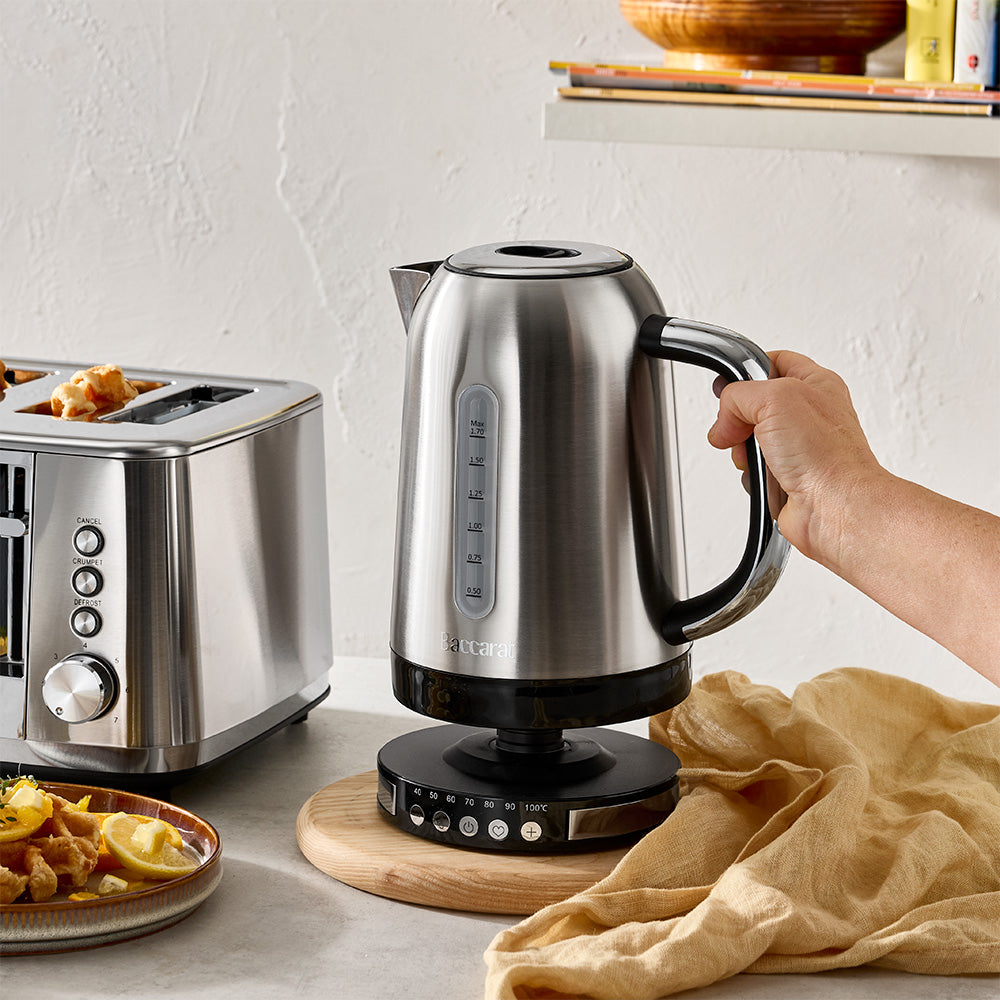 A stainless steel electric kettle with a black handle and base sits on a wooden trivet on a kitchen counter, with a hand holding the handle. A stainless steel toaster and a plate of toast are visible on the left, and a beige cloth is draped in the foreground.