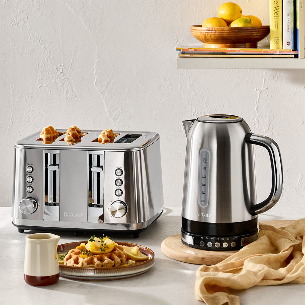 A stainless steel electric kettle and a four-slice toaster are placed together on a wooden board next to a plate of waffles.