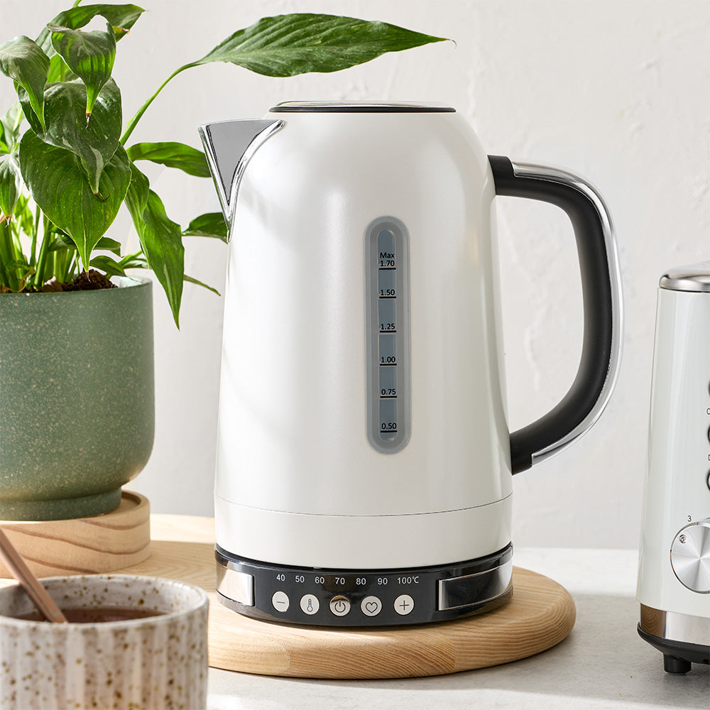 A white electric kettle is set on a wooden coaster on a countertop next to a potted plant.