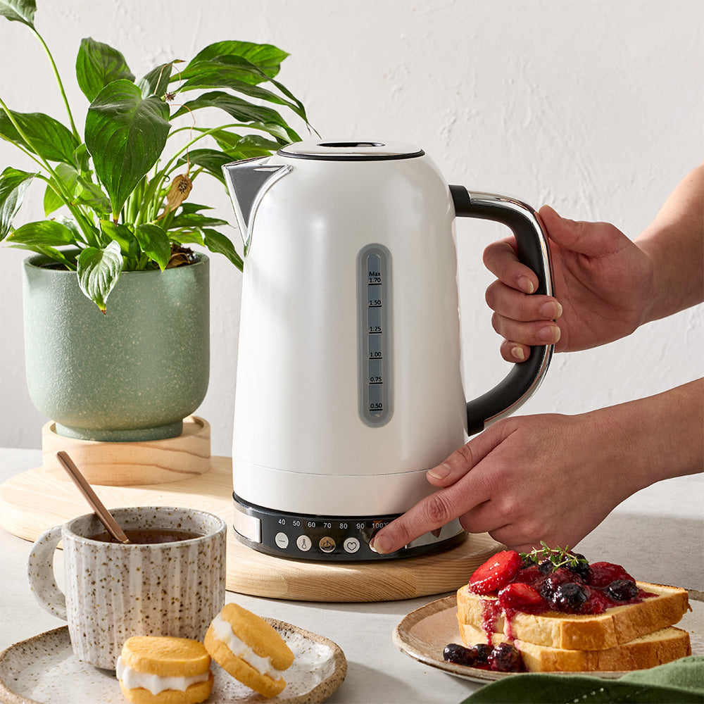 A person's hand is pressing a button on the base of a white electric kettle placed on a kitchen counter next to snacks.