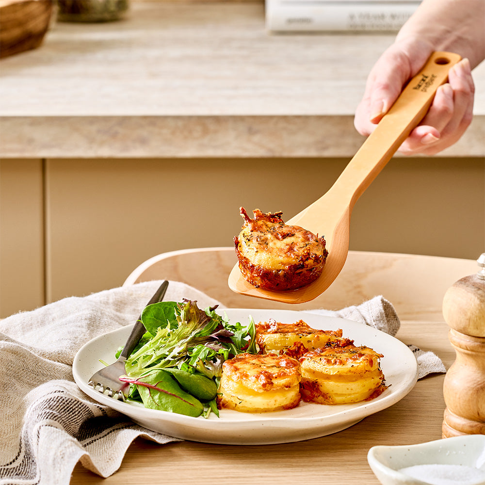 A hand uses the light brown wood spatula spoon to serve a portion of food onto a plate containing cooked scallops and a fresh side salad.