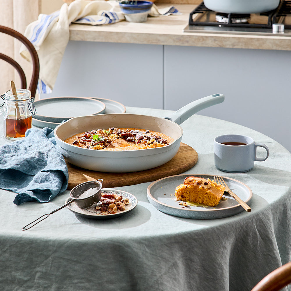 A light blue frying pan filled with cooked food sits on a wooden trivet on a table with a meal set out.