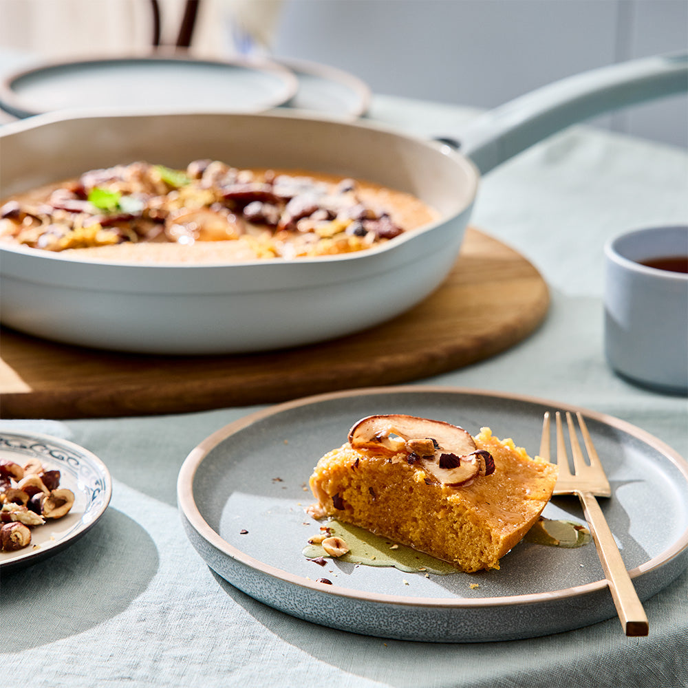 A slice of cake is served on a small plate next to a light blue frying pan containing the remaining portion.