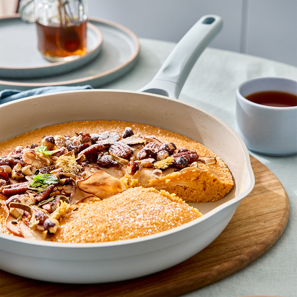 A white non-stick skillet filled with a savory baked dish topped with nuts, served on a wooden board at a dining table with plates and tea in the background.
