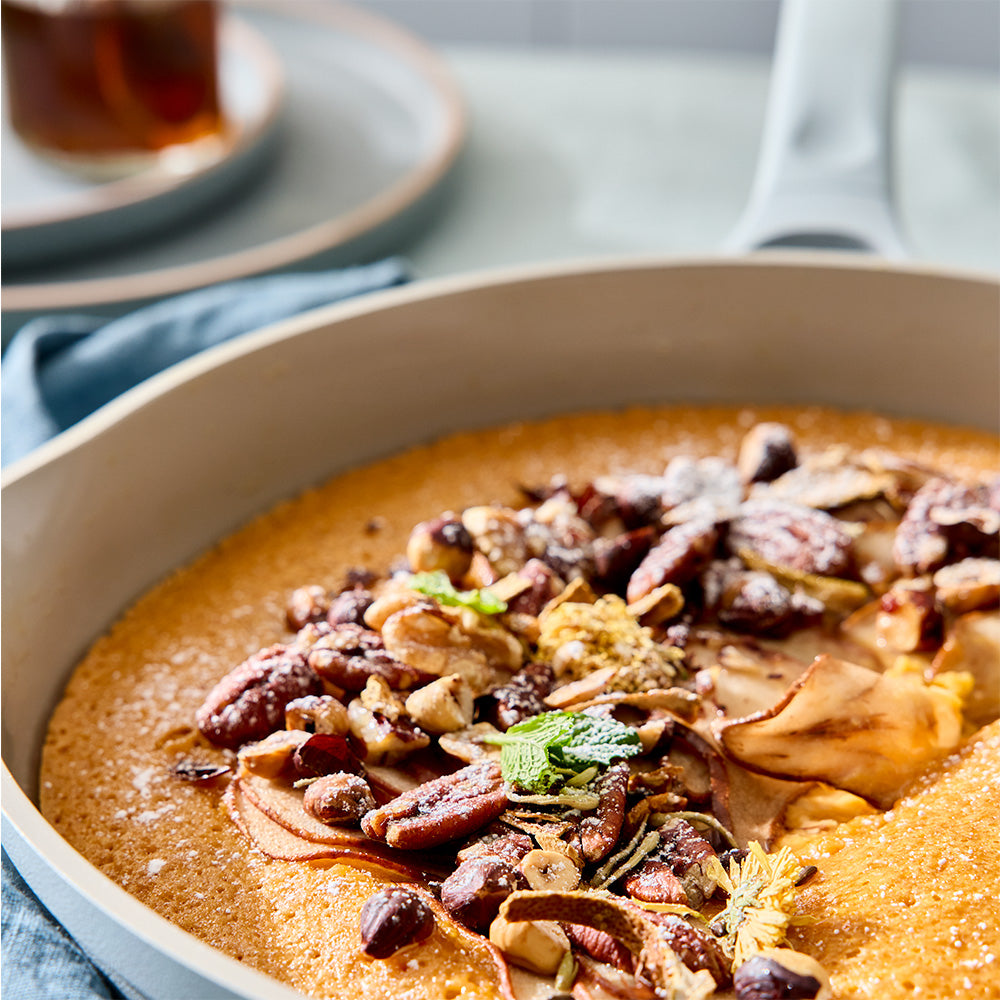 A close-up of a baked pumpkin pie in a light beige skillet, topped with chopped pecans, sliced apples, and a small mint garnish, set on a table with a blurred background.