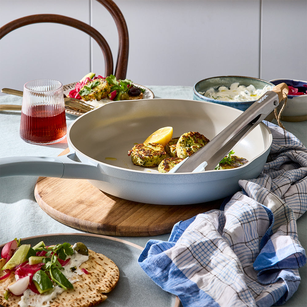 A light blue skillet with a serving utensil inside is set on a wooden board next to a meal and drinks.