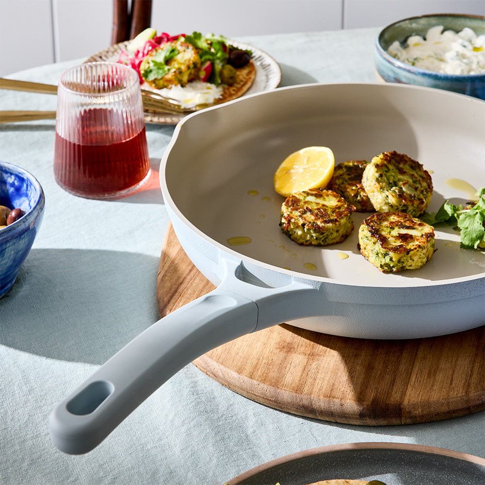 A light blue skillet containing cooked food and lemon slices is displayed on a dining table next to drinks and side dishes.