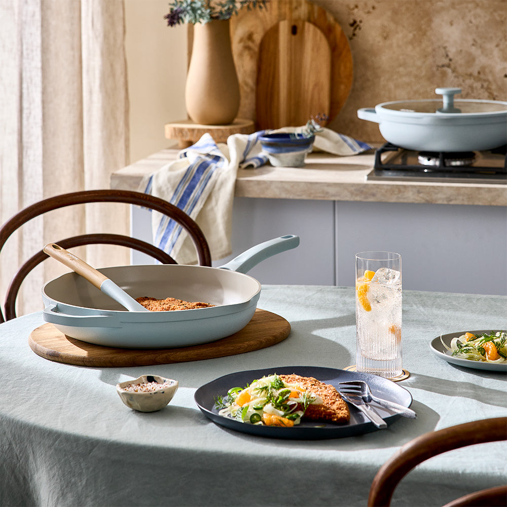 A light blue frying pan is displayed on a table next to a served meal, drink, and kitchen countertop setup.