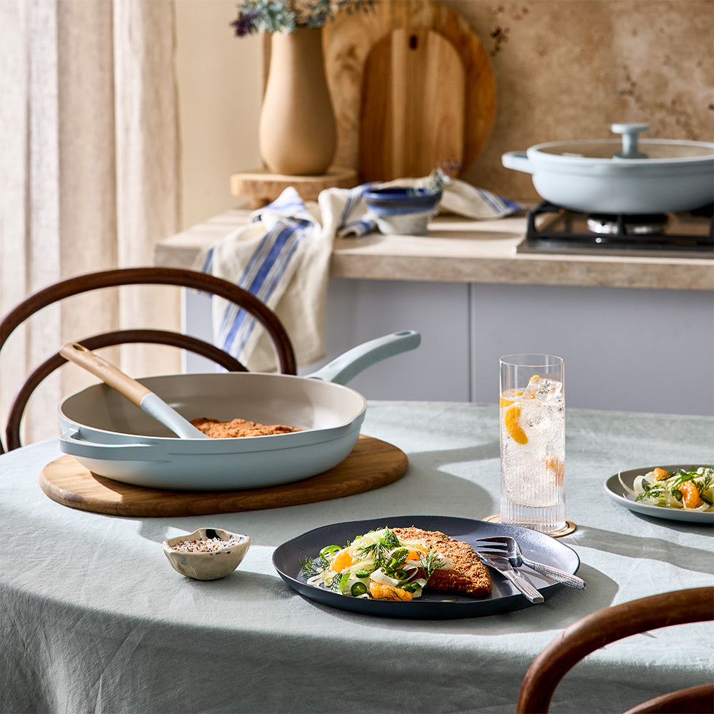 A light blue frying pan is set on a dining table alongside a served dinner plate, drinks, and kitchen decor in the background.