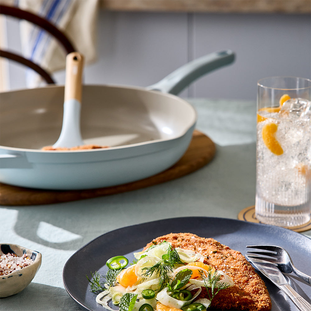 A light blue frying pan containing food is placed on a table next to a served plate of food and a cold drink.