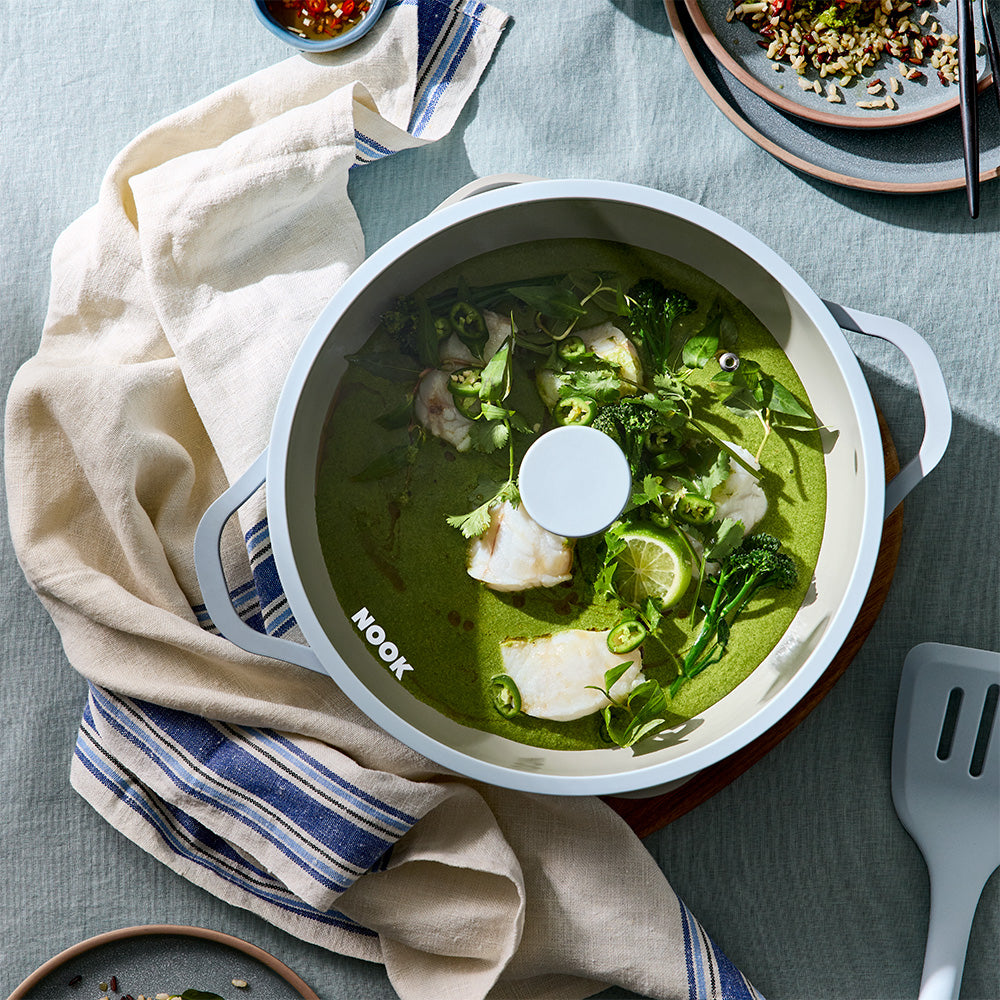 A Nook brand white ceramic pot filled with a green curry soup, shown from a top-down angle on a textured blue-gray tablecloth with a beige linen napkin and cooking utensils.