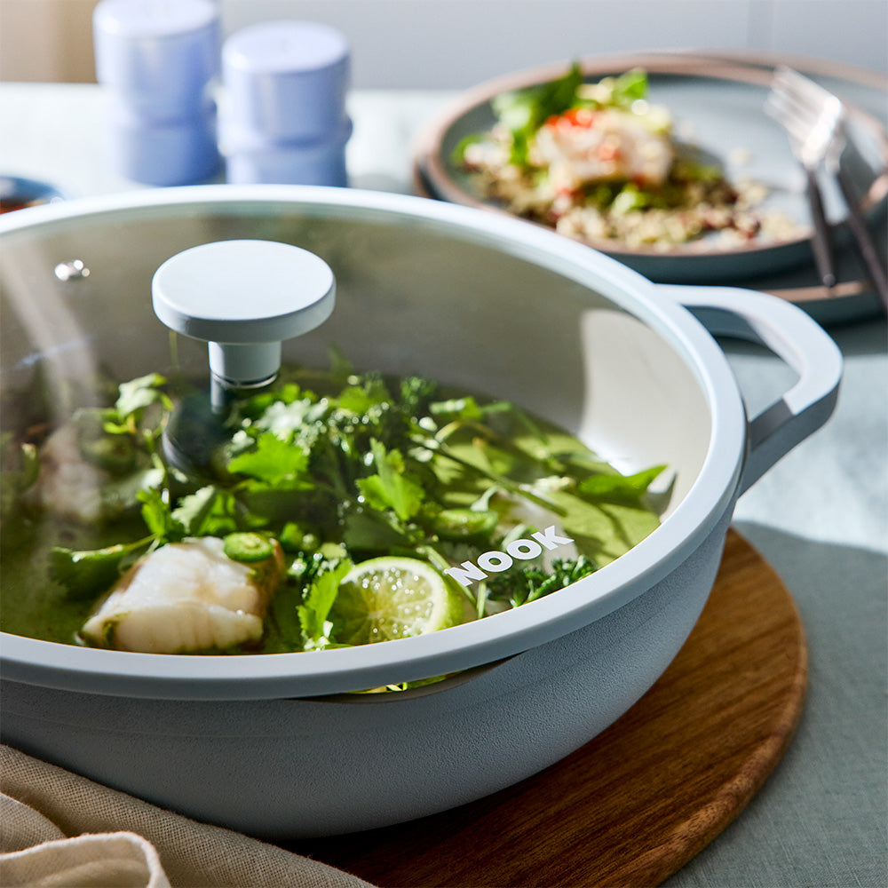 A light gray Nook brand ceramic pot with a clear glass lid, shown on a wooden board in a kitchen setting with food in the background.