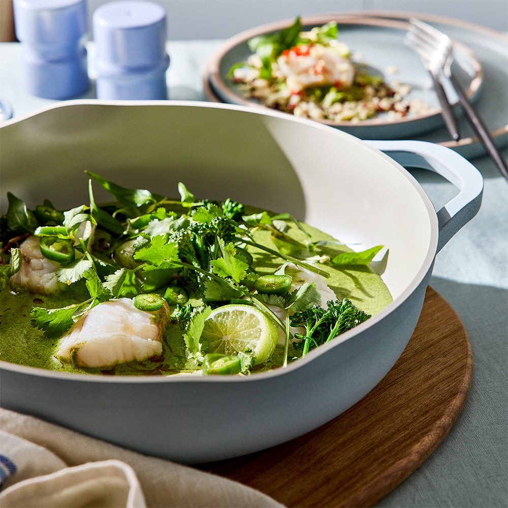 A light gray enameled cast iron skillet filled with a green fish and shrimp curry dish, garnished with cilantro and lime, set on a wooden board with a blurred plate of food in the background.