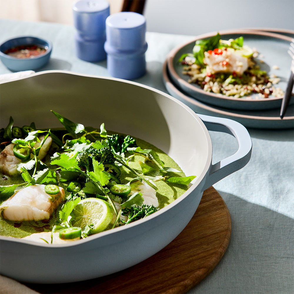 A light blue sauté pan containing a green broth dish with fish and herbs is placed on a wooden trivet.