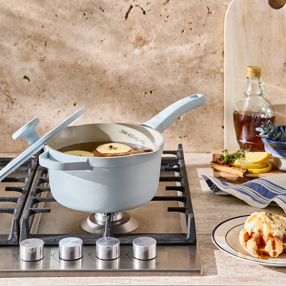 A light blue saucepan with its glass lid propped open sits on a gas cooktop near breakfast items.