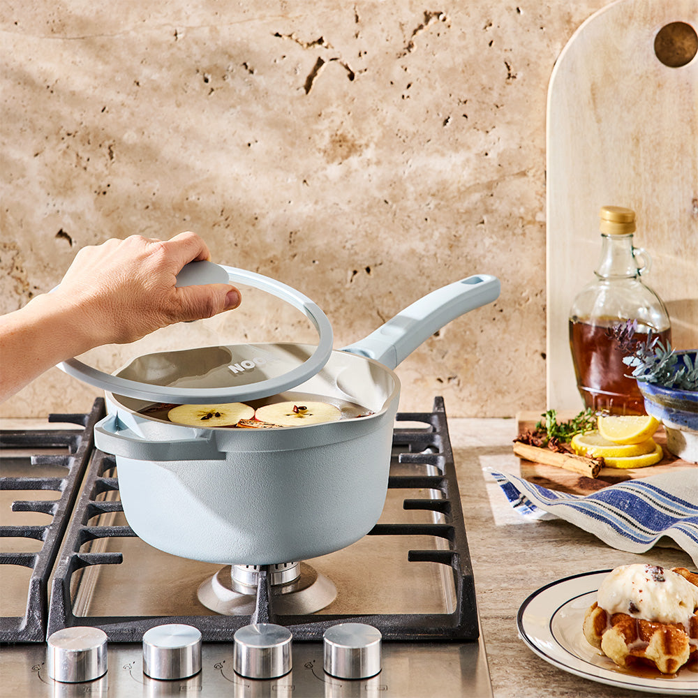 A person is lifting the glass lid off a light blue saucepan on a gas stovetop.