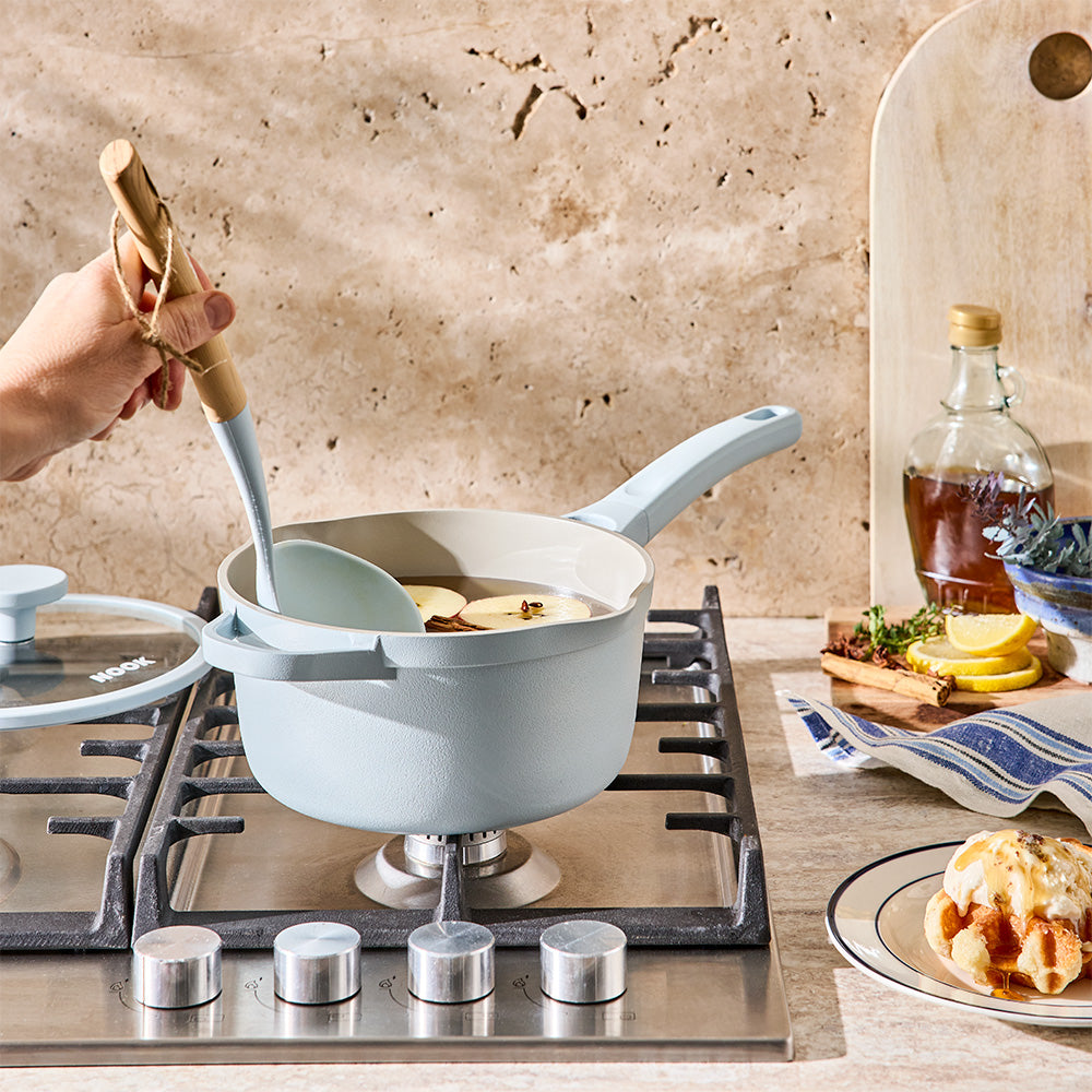 A person is using a wooden ladle to stir food inside a light blue saucepan on a cooktop.