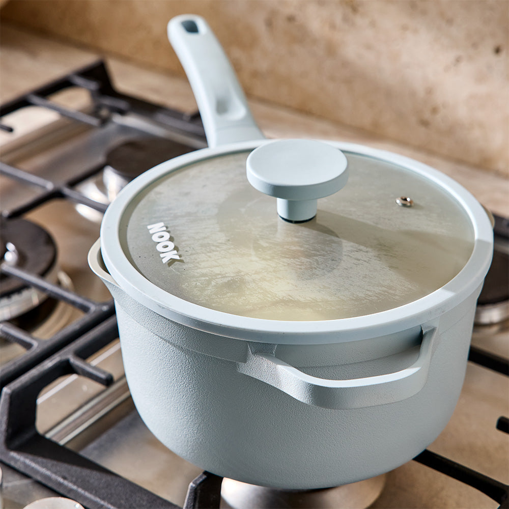 A light blue-gray enamel saucepan with a matching lid sits on a gas stovetop. The lid has a clear glass window and the brand name 'Nook' is printed on it.