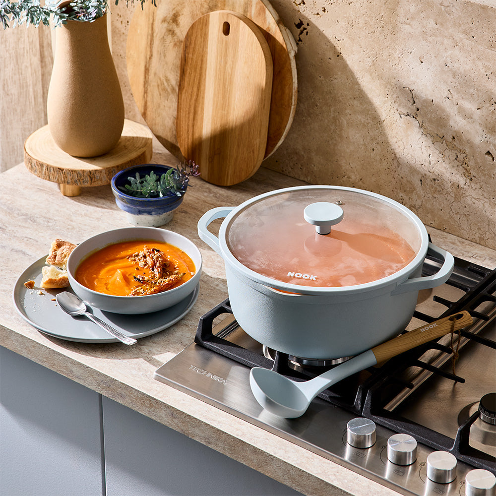 A light blue pot with a glass lid is placed on a stovetop burner next to a ladle and serving bowl.