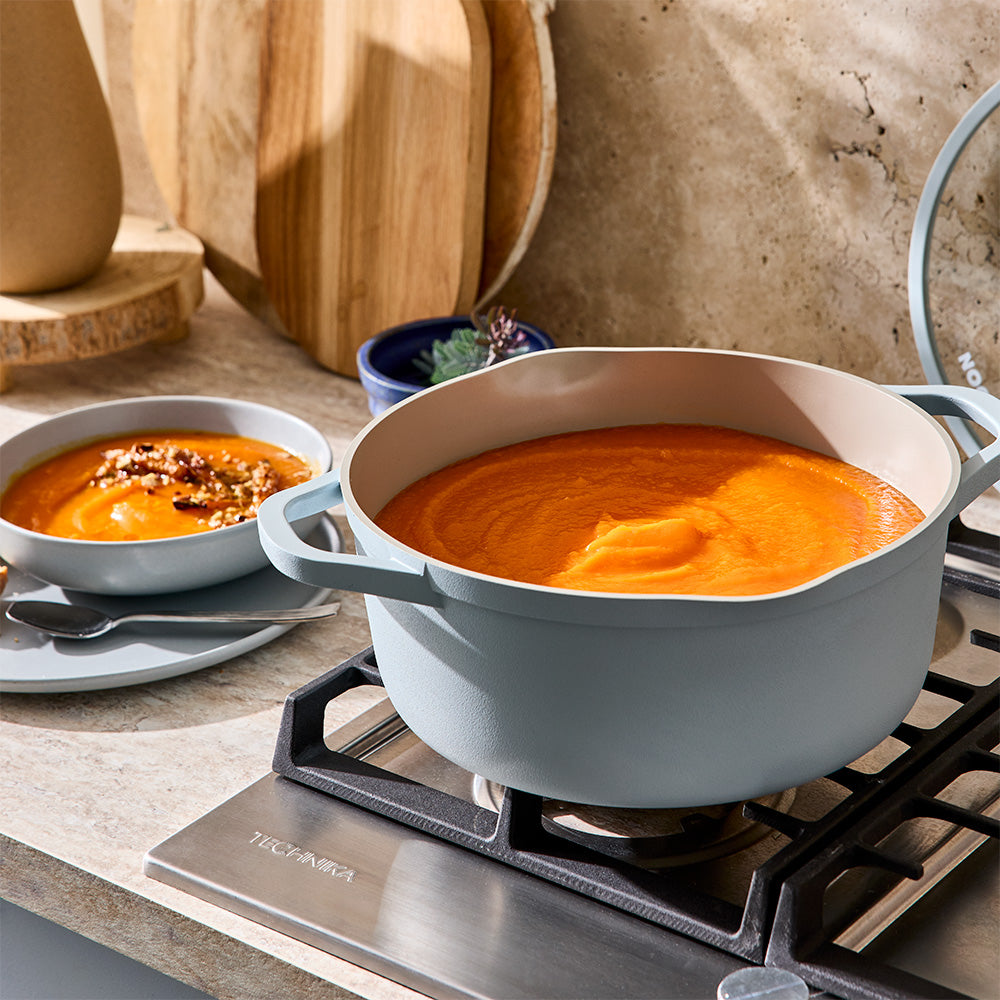 A light blue stockpot filled with orange soup is positioned on a stovetop next to a serving dish.