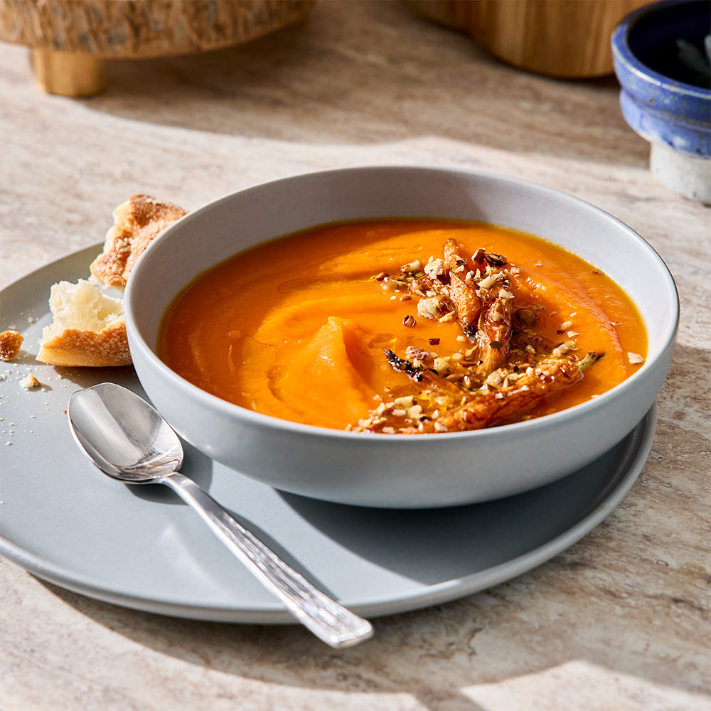 A bowl filled with orange soup is served on a placemat alongside a light blue cooking pot.