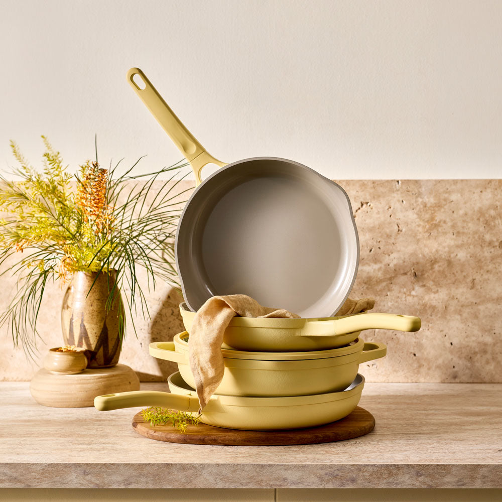 The NOOK by Baccarat 4 Piece Cookware Set Butter is stacked on a wooden board against a warm stone kitchen backdrop with a vase of dried flowers.