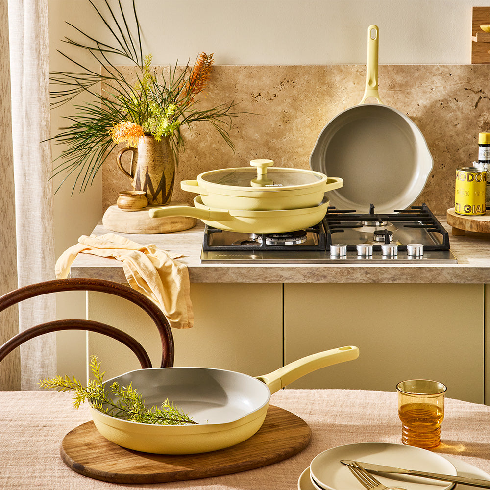 A pastel yellow skillet is set on a dining table alongside matching cookware on the kitchen counter in the background.