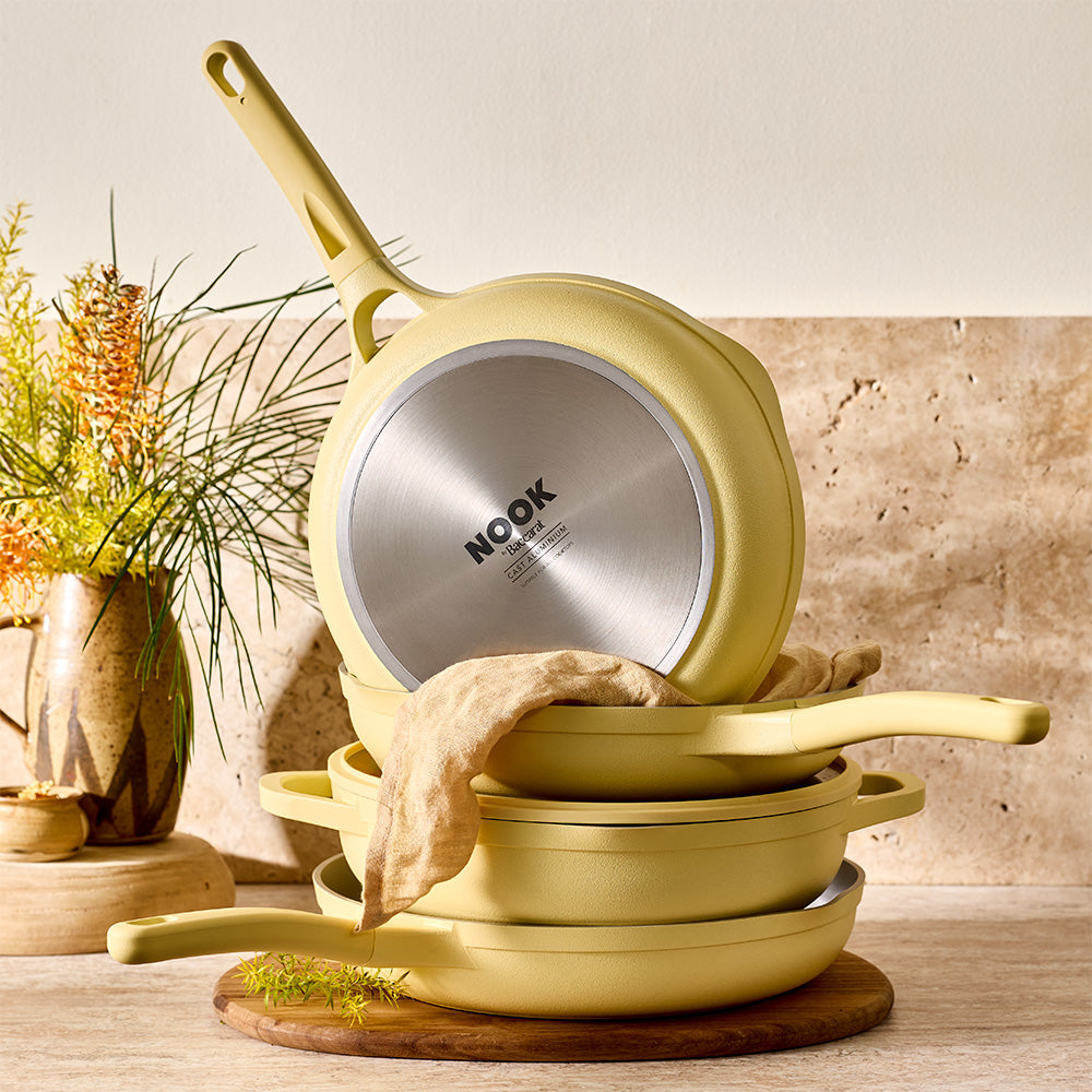 A stack of pastel yellow cookware is arranged on a wooden board next to a vase of flowers.