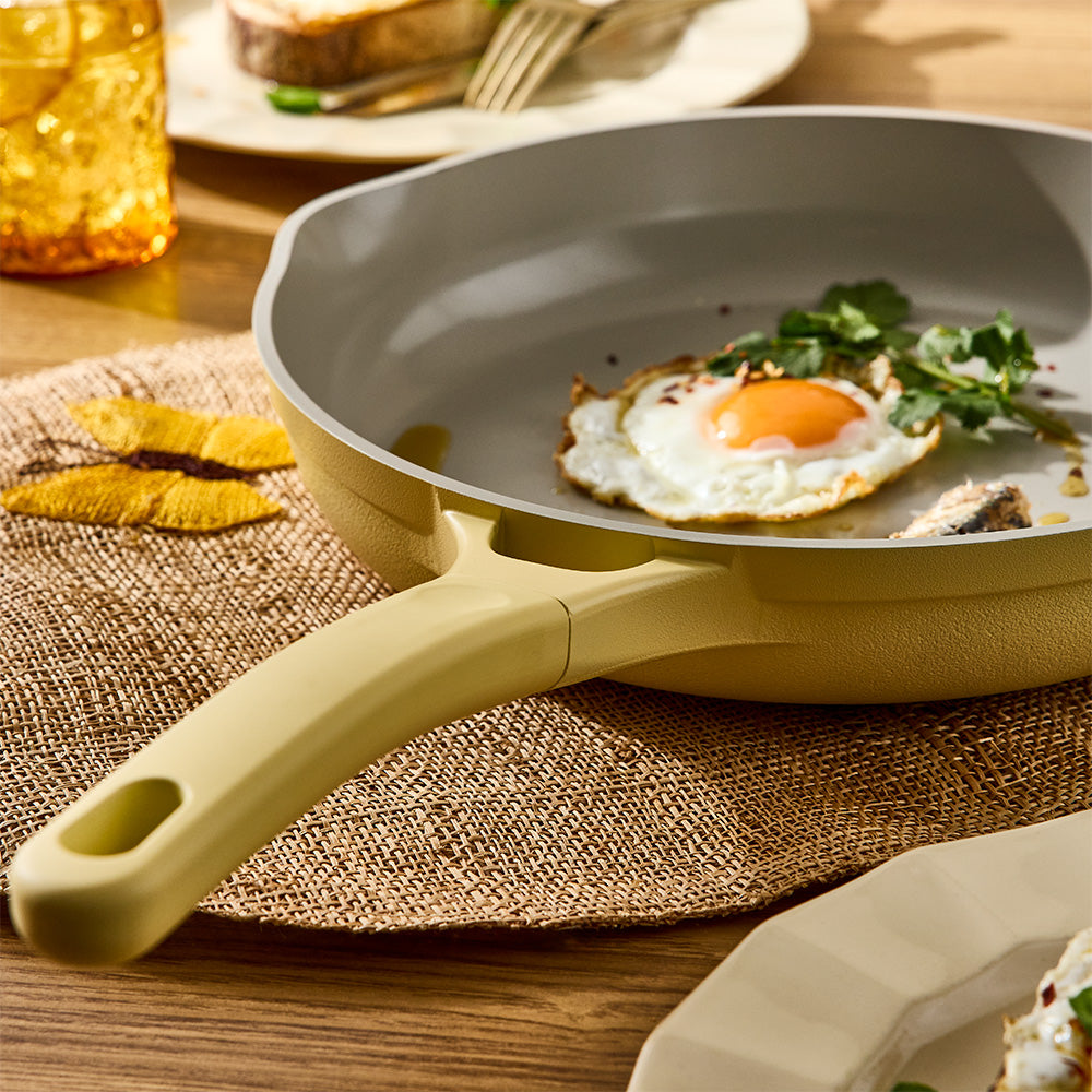 A light beige non-stick frying pan with a matching handle, shown on a woven placemat over a wooden table. A cooked egg with a bright orange yolk and green herbs are frying in the pan, with a plate and glass blurred in the background.