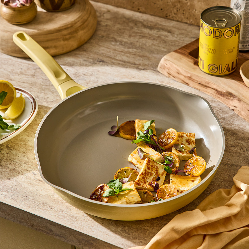 A pastel yellow skillet containing cooked food is set on a light-colored countertop next to cooking ingredients.