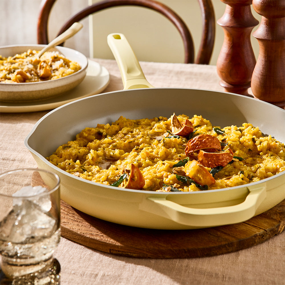 A pale yellow cast-iron skillet filled with a yellow rice dish, set on a wooden board on a dining table with a glass of water, a bowl of food, and wooden salt and pepper shakers in the background.