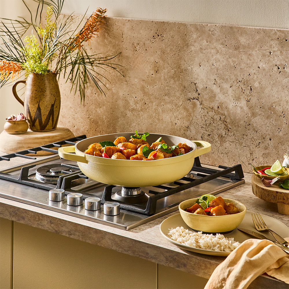 A pastel yellow sauté pan filled with cooked food sits on a gas stovetop in a warm kitchen setting.