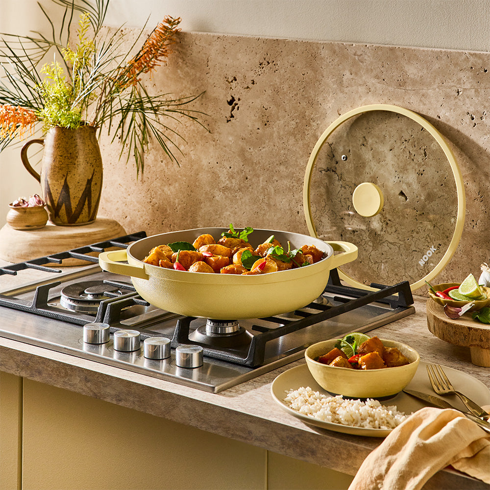 A pastel yellow sauté pan filled with food rests on a cooktop near a served bowl and side dishes.