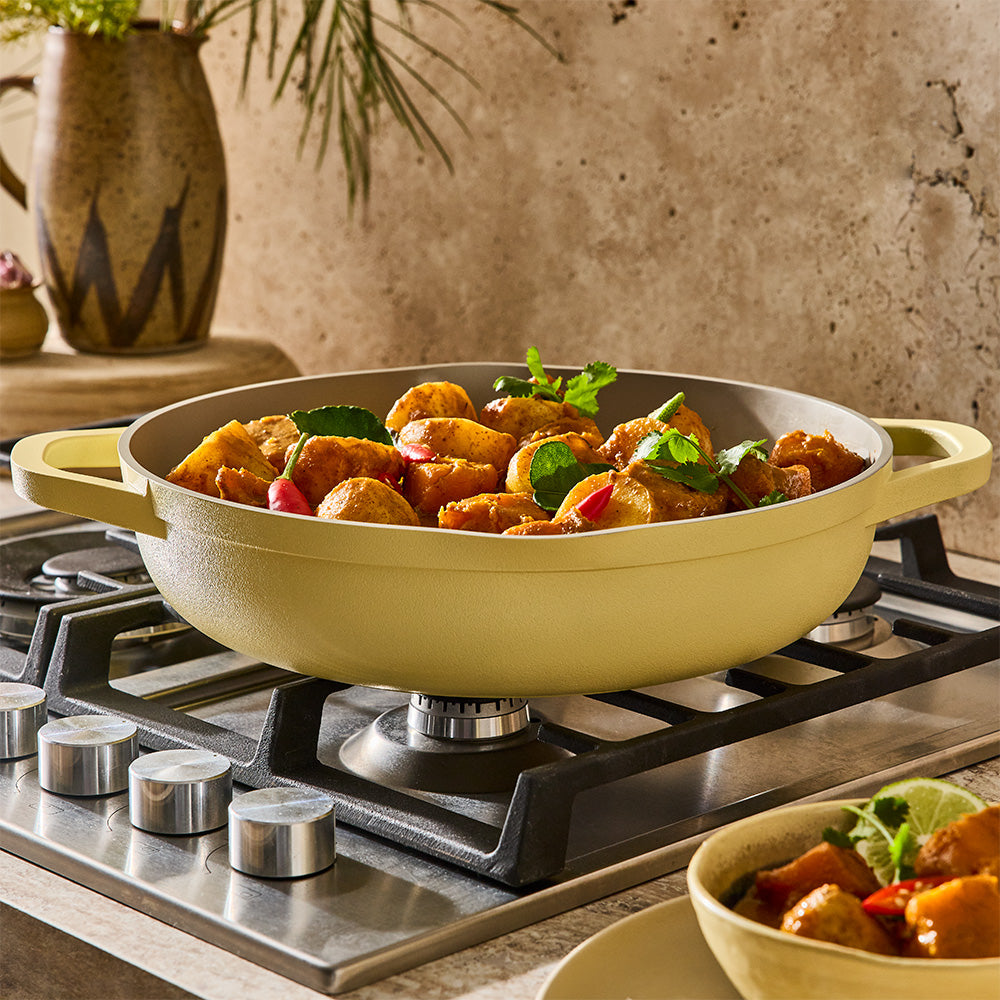 An uncovered pastel yellow sauté pan containing a cooked meat and vegetable dish sits on a cooktop.