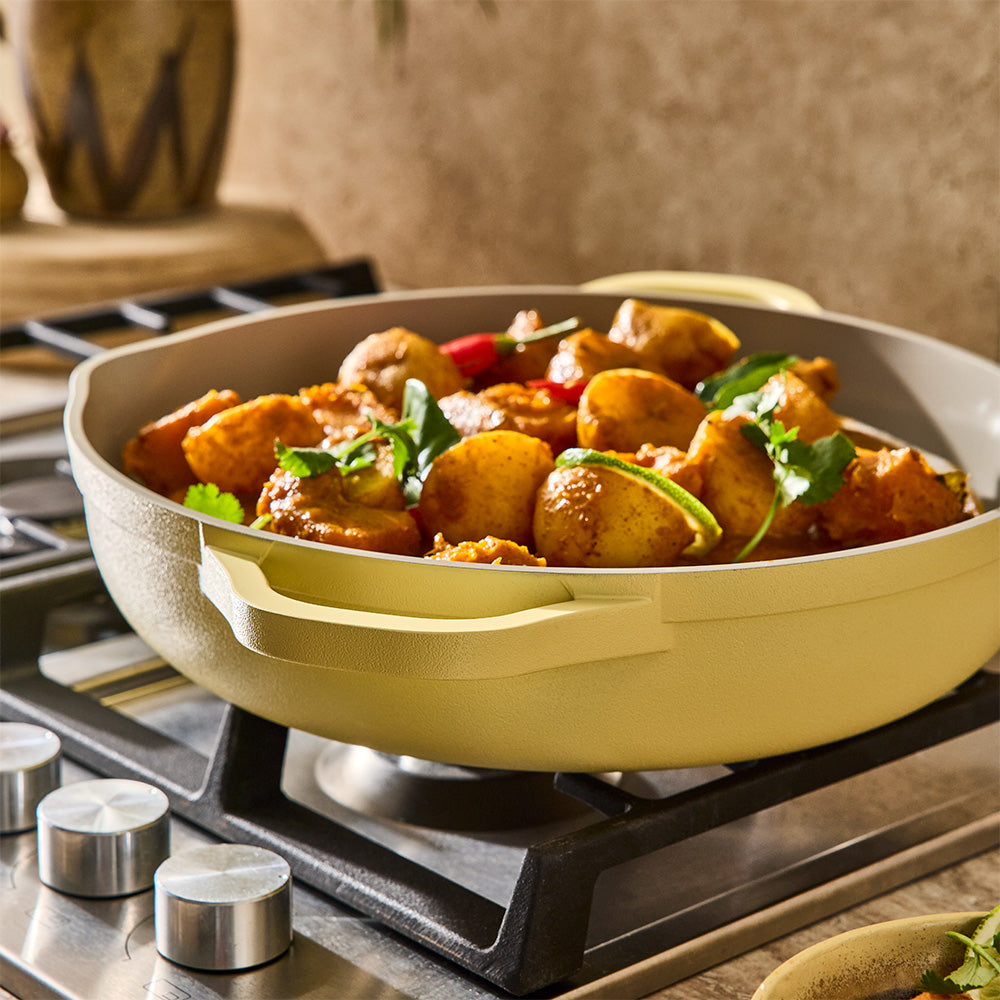 A pastel yellow sauté pan filled with cooked chicken and vegetables rests on a gas stovetop burner.