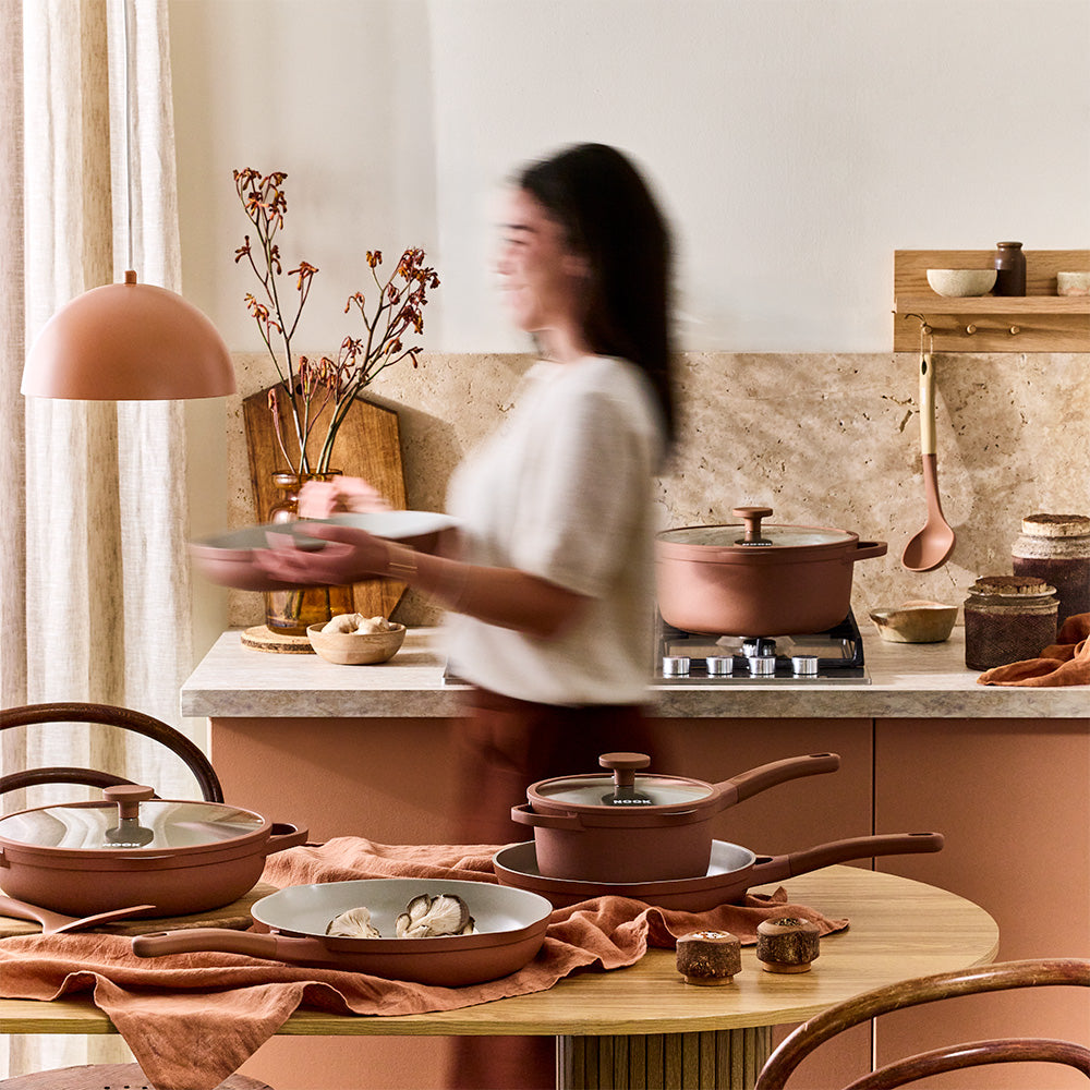 A woman in a white top and brown skirt carries a plate in a warm, rustic kitchen setting, with terracotta cookware and copper-toned cookware displayed on a wooden table and countertop.