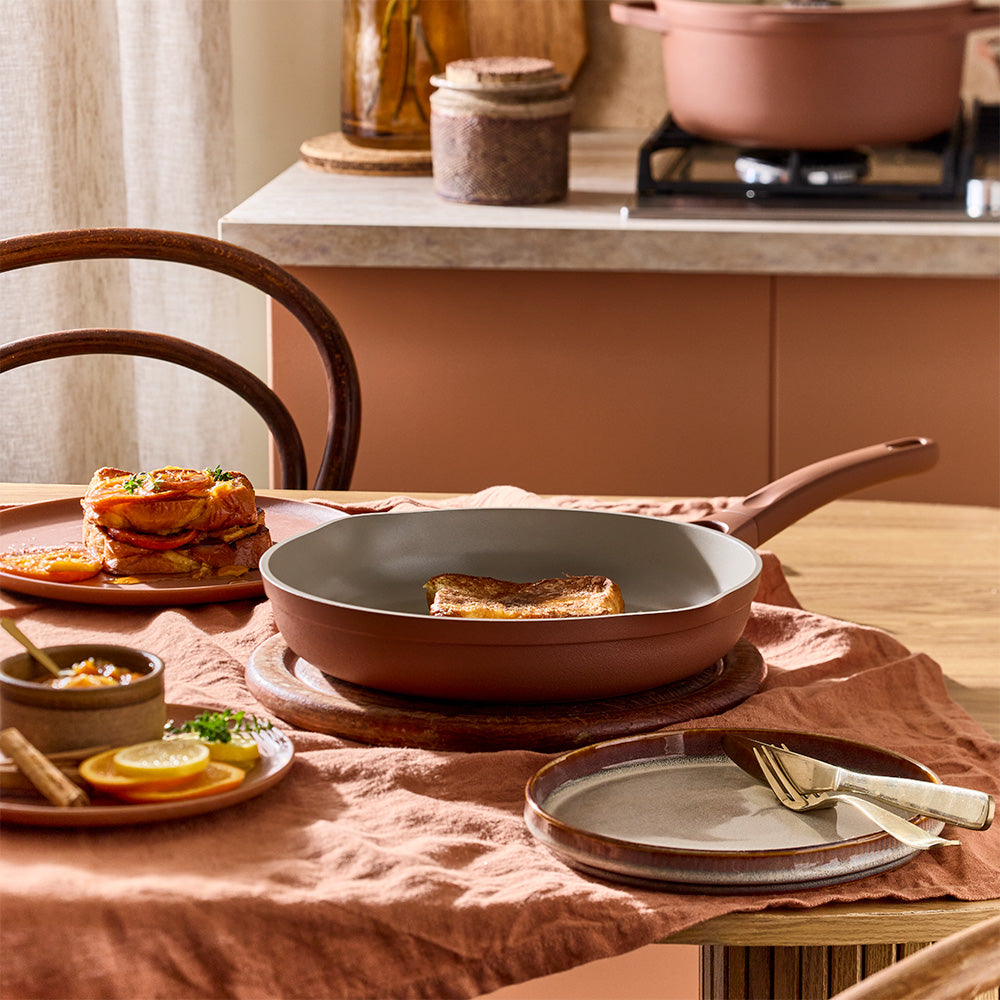 A terracotta-colored frying pan containing food rests on a dining table alongside served plates, cutlery, and side dishes.