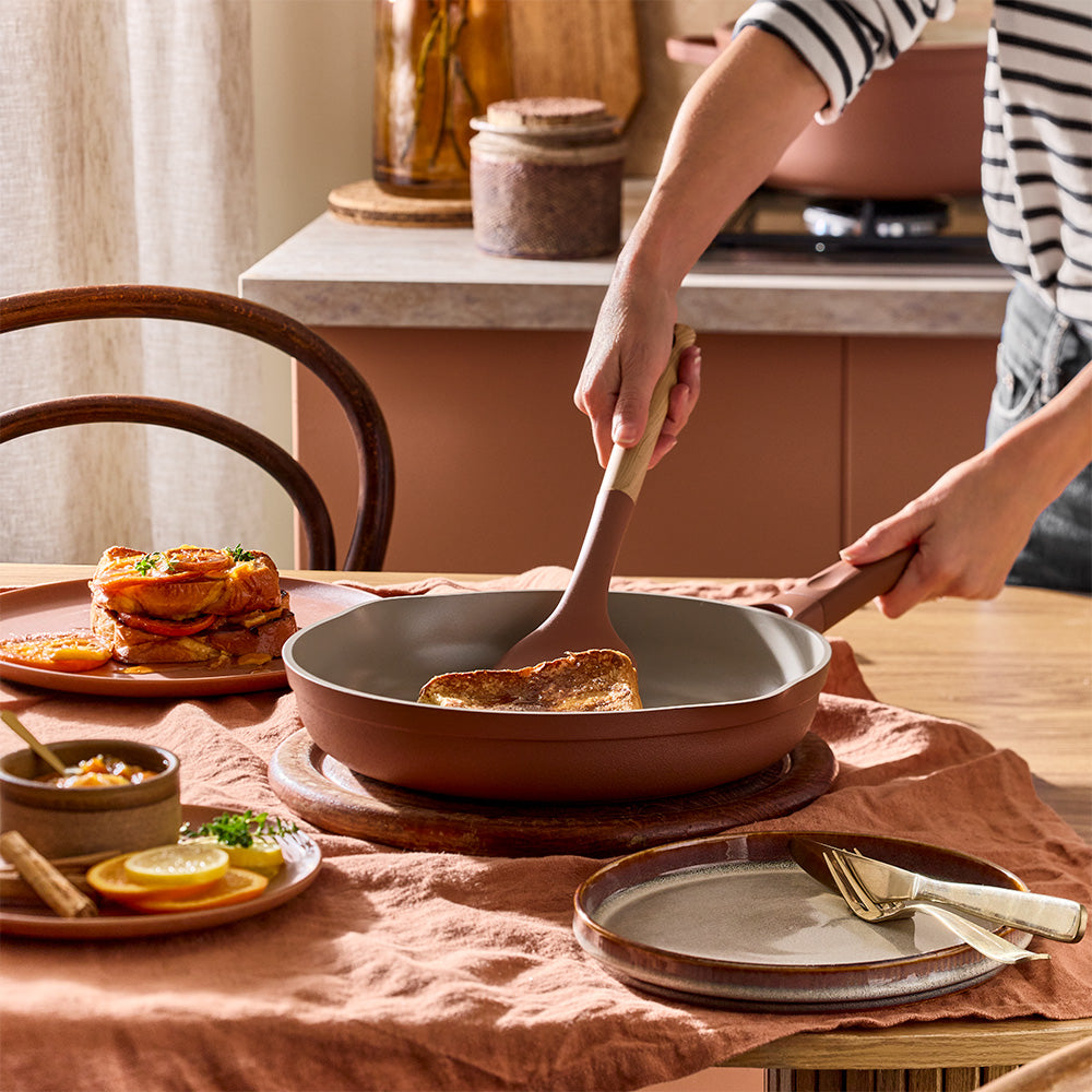 A person is using a spatula to stir food inside a terracotta-colored frying pan on a table set with a peach tablecloth.