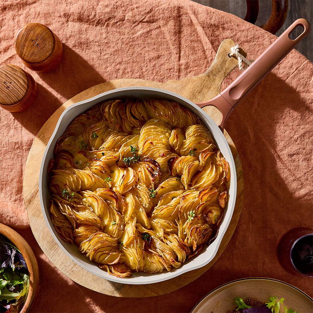 A cast iron skillet with a rose gold handle sits on a wooden board, filled with baked potato wedges topped with melted cheese and fresh chives, set on a rustic terracotta tablecloth.
