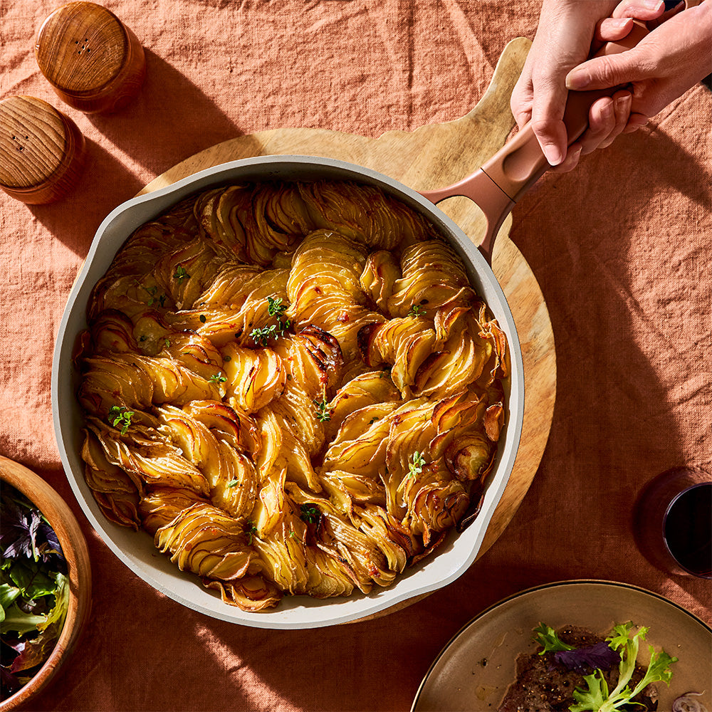 A top-down shot shows a person holding the handle of a blush pink skillet filled with baked potatoes on a peach tablecloth.