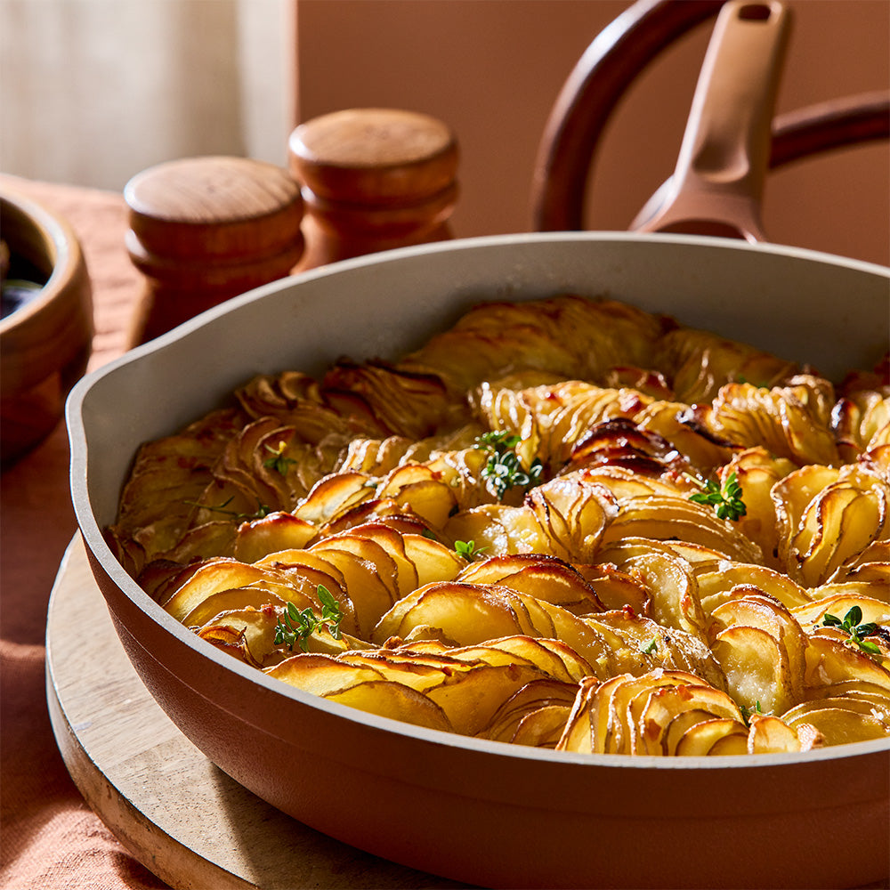 A close-up of a baked dish of layered potato slices with herbs, served in a brown ceramic skillet on a wooden board in a warm kitchen setting.